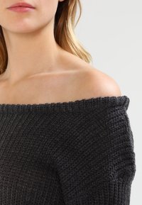 Pull en maille gris à épaules dénudées avec texture côtelée, encolure épaisse et coupe ample. Matière douce avec un design décontracté.