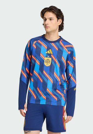 Ung mand iført en blå spansk fodboldtrøje med røde og gule diagonale striber og marineblå shorts.