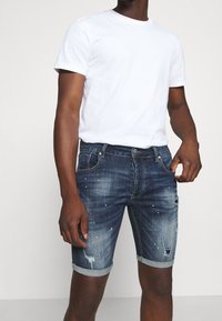 Pantalones cortos de mezclilla de color azul oscuro con un diseño desgastado y puños enrollados. Presentan un ligero desvanecimiento y adornos, combinados con una camiseta blanca básica.
