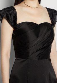 Černé saténové šaty s výstřihem do srdce, které mají draped úpravy na těle a krátké puff rukávy. Hladká textura s minimálním leskem.