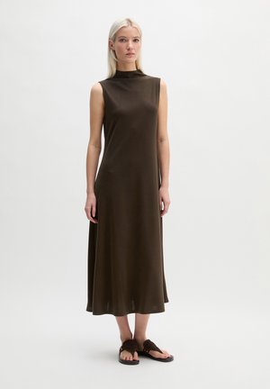 Kobieta w ciemnobrązowej sukience midi bez rękawów i ciemnobrązowych sandałach stojąca na tle jednolitego jasnego tła.