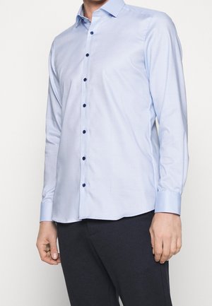 Man wearing a light blue long-sleeve button-up shirt with dark blue buttons and dark pants against a plain background.