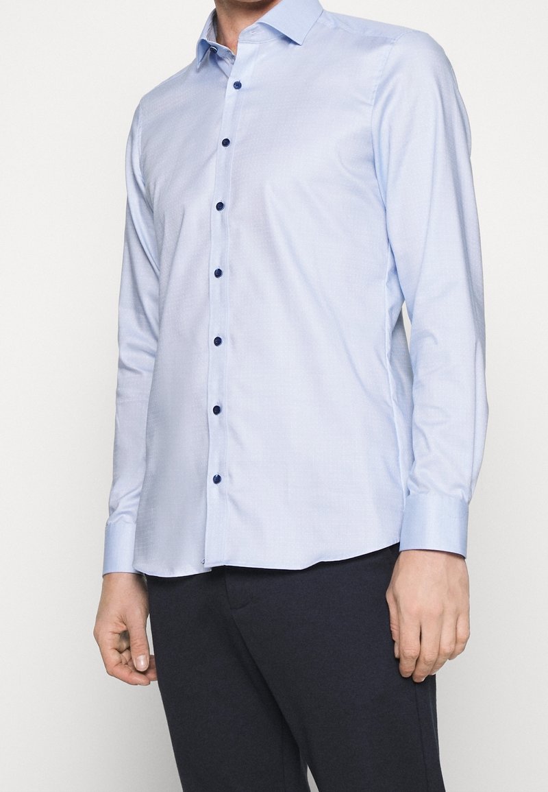 Homme portant une chemise bleu clair à manches longues avec des boutons bleu foncé et un pantalon foncé sur un fond uni.