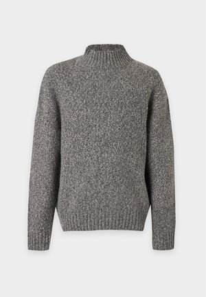 Jeune homme portant un pull en laine gris, un pantalon beige à carreaux, des bottes noires et une ceinture, debout devant un fond blanc uni.