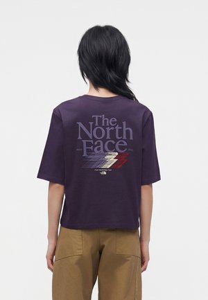 Personne aux cheveux noirs portant un t-shirt The North Face violet foncé avec un texte graphique dans le dos, et un pantalon marron clair, debout, dos à l’objectif.