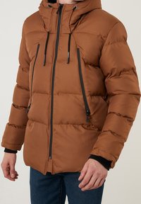 Brun pufferjacka med huva, har dragkedja framtill, svarta detaljer och ett quiltat mönster. Inkluderar dragsko på huvan.