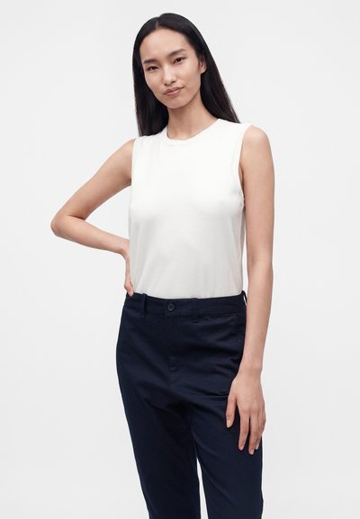 Jeune femme aux longs cheveux foncés portant un haut blanc sans manches et un pantalon bleu marine foncé, debout avec une main sur la hanche.