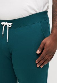 Teal joggingbroek met een elastische tailleband, trektouwsluiting en voortassen. De stof lijkt zacht en flexibel.