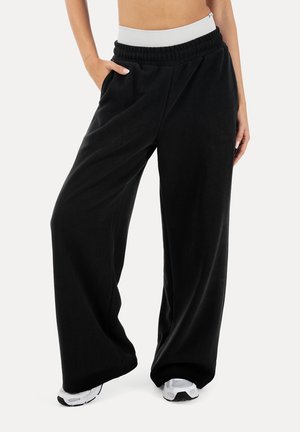 Schwarze Sweatpants mit weitem Bein, elastischem Bund und Seitentaschen, getragen mit weißen Sneakers und einem darunter sichtbaren weißen Bund.