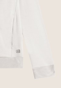 Sweatshirt blanc avec un tissu texturé, des poignets côtelés, une poche latérale et un petit accent en métal sur le côté gauche.