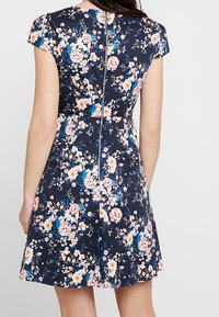 Femme portant une robe bleu marine à manches courtes avec des motifs floraux roses et blancs, et une fermeture éclair argentée visible le long du dos.