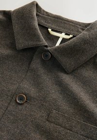 Brown textured polo shirt featuring a pointed collar, two dark buttons, and a chest pocket with a subtle woven pattern.