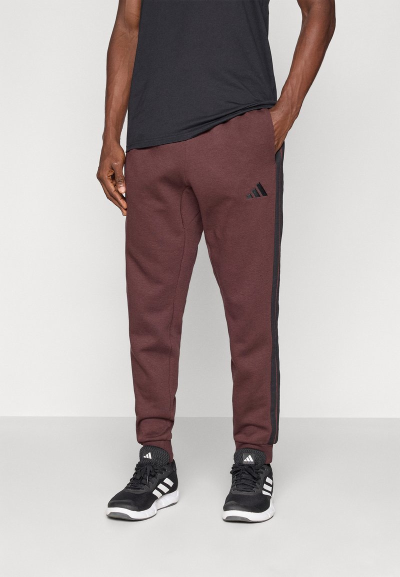 Bordo sporta bikses ar melnām sānu svītrām un izšūtu melnu Adidas logo augšstilbā. Valkātas ar melniem sporta apaviem.