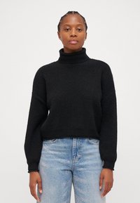 Maglione nero a collo alto con un design a maglia testurizzata, caratterizzato da maniche ampie e una vestibilità rilassata, abbinato a jeans azzurri chiari con decorazioni.