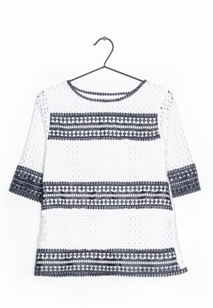 Witte semi-doorzichtige blouse met zwarte geometrische geborduurde strepen op mouwen, borst en zoom, hangend aan een zwarte metalen hanger.