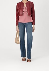 Rote Lederjacke in Cropped-Form über einem rosa Top mit Knoten-Detail, kombiniert mit blauen Jeans und braunen, gesteppten Sandalen. Hält eine beige Handtasche.