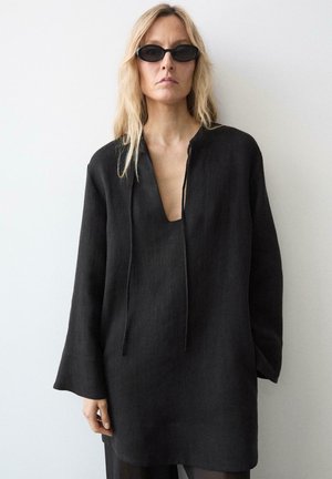 Blonde Frau trägt eine schwarze, übergroße Tunika mit tiefem V-Ausschnitt und schwarze, schlanke Sonnenbrille, steht vor einer schlichten hellen Wand.