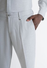 Pantaloni grigi chiari su misura realizzati in tessuto testurizzato, con chiusura a bottone, vita regolabile e tasche laterali.