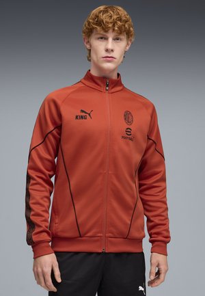 Giacca atletica rossa con cerniera frontale, colletto alto, dettagli neri e logo sul petto; presenta strisce con motivo sulle maniche.