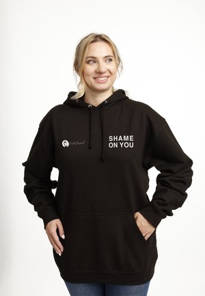 Smilende kvinde iført en sort hættetrøje med teksten "SHAME ON YOU" foran, stående med den ene hånd i lommen mod en hvid baggrund.