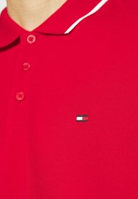 Polo rouge en tissu texturé, avec une patte de boutonnage à quatre boutons et un petit logo brodé en bleu, blanc et noir sur la poitrine.