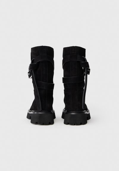 Par de botas de ante negras hasta la mitad de la pantorrilla con suelas gruesas y decorativas correas en los tobillos, vistas desde atrás sobre un fondo liso.