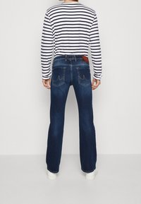 Jeans oscuros de mezclilla con un diseño de pierna recta, presentando una etiqueta de cuero marrón en la cinturilla y bolsillos traseros visibles. Camisa de rayas de manga larga en la parte superior.