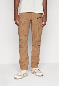 Khaki cargobroek van katoenmix met meerdere zakken, een met rits afgesloten zijzak en een taps toelopend ontwerp. G gedragen met witte sneakers.