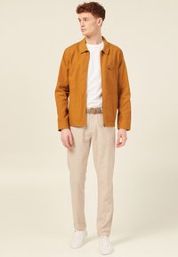 Brun vävd jacka, vit t-shirt, beige byxor och vita sneakers. Jackan har krage och bröstficka. Avslappnad outfit för varmt väder.