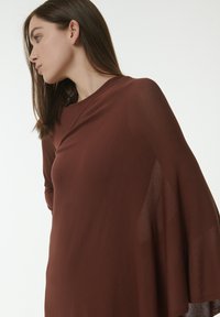 Haut en tissu léger marron avec une coupe loose et des manches larges. Texture douce, design minimaliste, drape élégamment autour du corps.