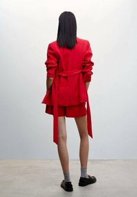 Blazer rojo a medida con cinturón ajustable, combinado con pantalones cortos a juego. Cuenta con mangas largas y un diseño en capas. Zapatos negros y calcetines grises.