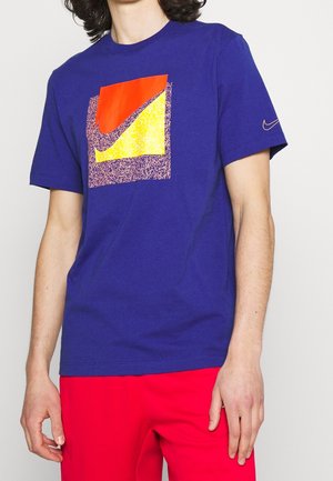 Camiseta de algodón azul royal con un diseño gráfico multicolor que tiene acentos en rojo, amarillo y moteados. Logo de Nike en la manga.