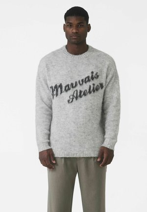 Jasnoszary sweter dziany o puszystej fakturze z napisem „Mauvais Atelier” w ciemnoszarym stylu pisma na przodzie.