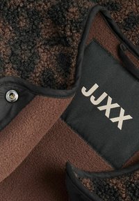 Smeđi flis materijal s teksturiranim, tamnim uzorkom prekriva sivi panel na kojem se nalazi logo "JJXX" u svijetlosivoj boji. Vidljiv metalni detalj s kopčom.