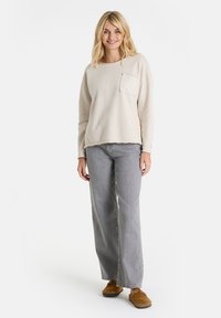 Lächelnde Frau mit blonden Haaren trägt ein beiges Langarmoberteil, graue weit geschnittene Jeans und braune Schuhe, steht vor einem weißen Hintergrund.