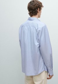 Jonge man met krullend haar, gekleed in een lichtblauw gestreept shirt met lange mouwen en crèmekleurige broek, met zijn rug naar de kijker tegen een effen achtergrond.