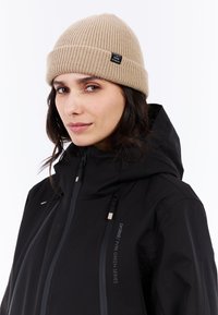 Zwarte waterdichte jas met een hoge kraag, voorzien van grijze ritsen. Draagt een beige, geribbelde gebreide beanie met een logo-label aan de zijkant.