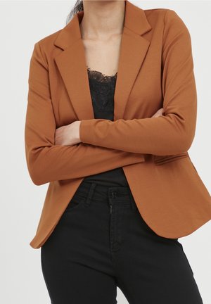 Kobieta w rdzawej marynarce na czarnej bluzce z koronkowym wykończeniem, z założonymi skrzyżowanymi ramionami, w połączeniu z czarnymi spodniami.