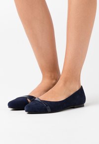 Navyblå semskede ballettsko med en blank bånd over tåen, rund form, og flat såle. Glatt tekstur og minimalistisk design.