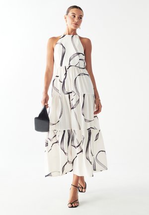Halter neck maxi dress in white with black wave patterns, tiered design, and lightweight fabric. Paired with a small black handbag and sandals.