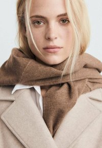 Écharpe marron enroulée autour du cou, fabriquée en tissu doux. Manteau beige clair avec un col accentué et une chemise blanche en dessous. Éclairage naturel.