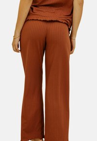 Pantalons côtelés de couleur rouille avec un design évasé. Tissu doux et texturé avec un ourlet à bords festonnés. Détails de couture visibles et grandes ouvertures pour les jambes.