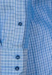 Niebieska koszula w kratę z wzorami w jasnoniebieskie i ciemnoniebieskie kratki. Posiada niebieskie guziki i fakturowaną tkaninę. Widoczny detal mankietu.