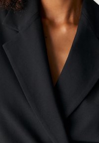 Blazer noir avec revers, présentant une coupe cintrée et une texture lisse. Le col visible et le décolleté en V créent une apparence structurée.