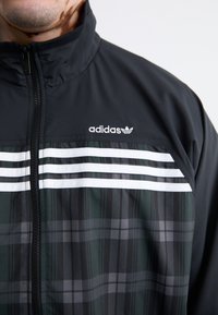 Juodas sportinis džemperis su užtrauktuku priekyje, su baltais dryžiais ir tamsiai žaliu bei pilku languotu raštu. Logotipas rodomas viršutiniame kairiajame kampe.