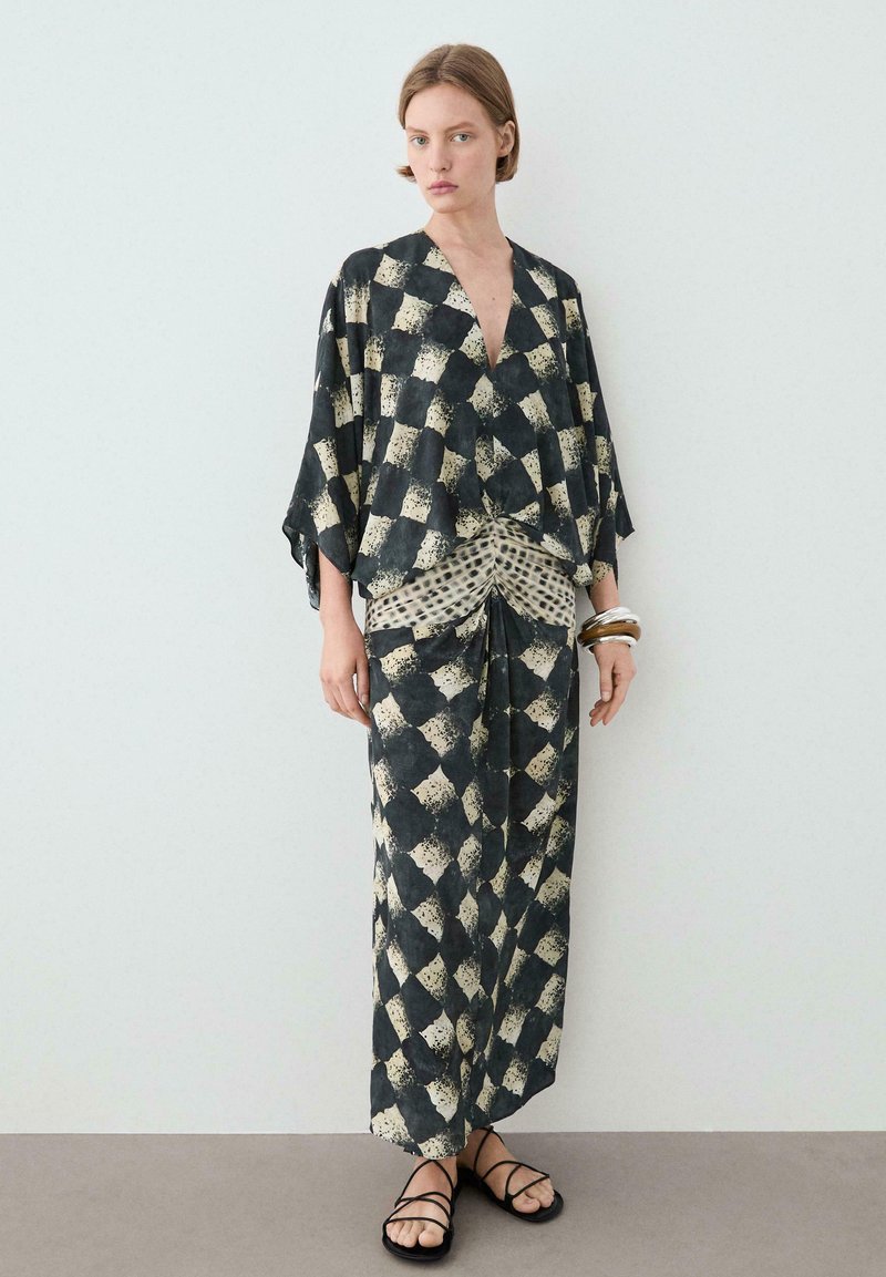 Frau trägt ein schwarz-beiges, abstrakt kariertes Kimono-Top und einen passenden langen Rock mit Rüschen-Detail, schwarze Sandalen und Armreifen.