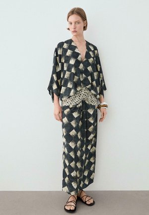 Frau trägt ein schwarz-beiges, abstrakt kariertes Kimono-Top und einen passenden langen Rock mit Rüschen-Detail, schwarze Sandalen und Armreifen.