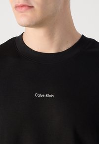 Black cotton t-shirt with a crew neckline featuring a white "Calvin Klein" logo on the left chest area, smooth texture.