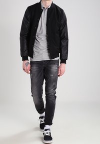 Veste bomber noire avec manches en cuir, polo gris, jean denim foncé avec détails usés, et baskets noires avec lacets blancs.