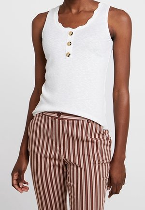 Débardeur blanc côtelé avec décolleté festonné et trois boutons dorés, associé à un pantalon rayé marron et crème.